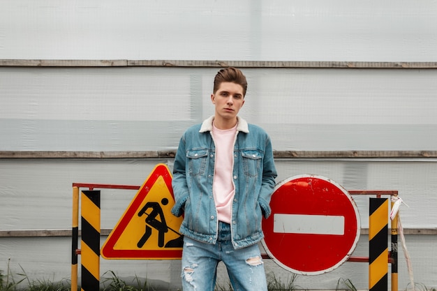 Knappe jonge stijlvolle man in trendy blauwe jeans vrijetijdskleding uit nieuwe collectie staan in de buurt van gebouwen en verkeersborden in de stad. Knappe jongen in modieuze denim kleding loopt op straat. Jeugd stijl.