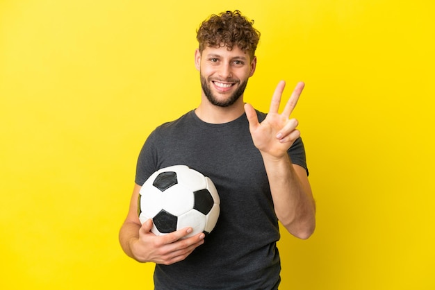 Knappe jonge voetballer man geïsoleerd op gele achtergrond gelukkig en drie tellen met vingers