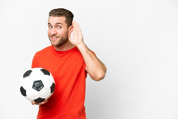 Knappe jonge voetballer man over geïsoleerde witte achtergrond luisteren naar iets door de hand op het oor te leggen