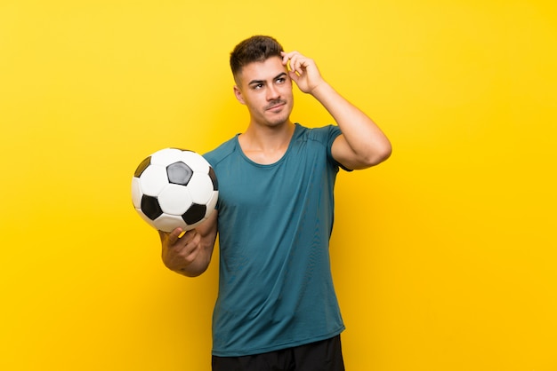 Knappe jonge voetbalstermens over geïsoleerde gele muur die twijfels hebben en met verwarren gezichtsuitdrukking