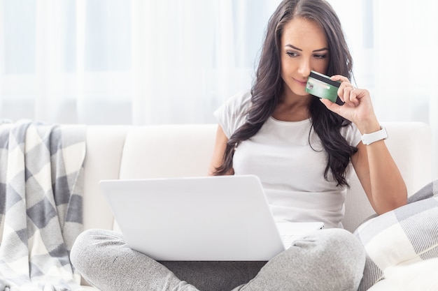 Knappe jonge vrouw overweegt online winkelen terwijl ze op haar witte bank zit met een laptop op haar schoot.