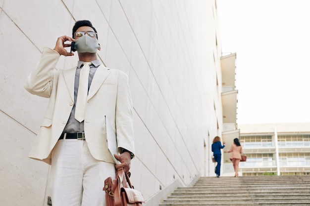 Knappe jonge zakenman in wit pak en medisch masker die de trap af loopt en het kantoorgebouw verlaat en telefoneert met een collega of zakenpartner