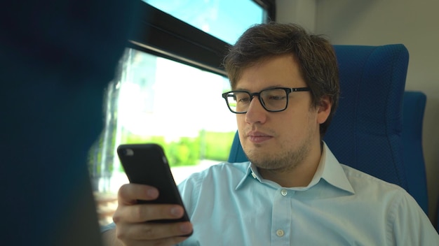Knappe jonge zakenman met een blauw shirt en bril rijdt in een trein die een nummer kiest en op zijn telefoon praat Zonnige zomerdag