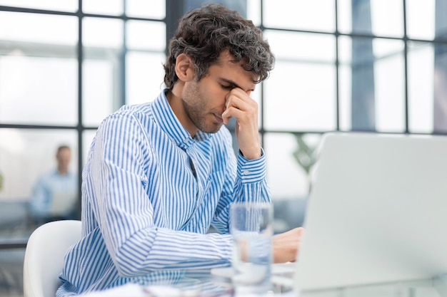 Knappe jonge zakenman met gesloten ogen die zijn gezicht met de handen aanraken terwijl hij op de werkplek op kantoor zit Collega is op de achtergrond