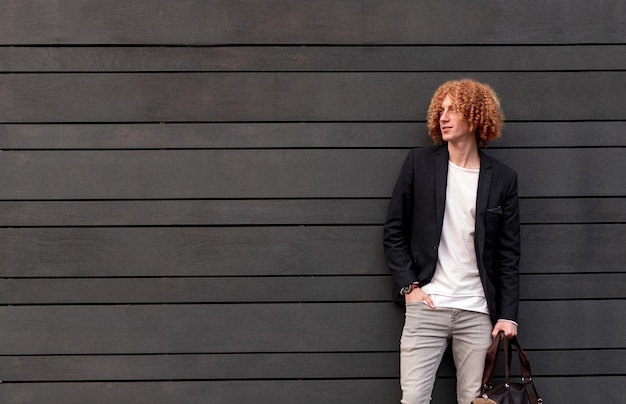 Knappe jonge zelfverzekerde man met rood krullend haar gekleed in trendy outfit met tas en wegkijken terwijl hij tegen een grijze muur staat