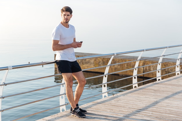 Knappe jongeman atleet die op de pier staat en smartphone gebruikt