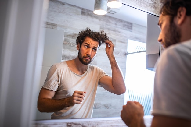 Knappe jongeman die zijn haar met de hand aanraakt en thuis in de badkamer verzorgt. blanke metroseksuele man maakte zich zorgen over haaruitval en keek naar de spiegel van zijn terugwijkende haarlijn.