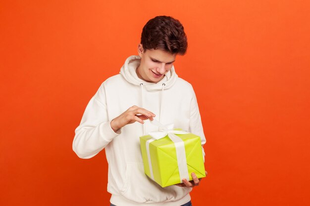 Knappe jongeman in casual wit sweatshirt met capuchon strik op geschenkdoos aanpassen en lachende tiener voorbereiden datum verjaardagscadeau Indoor studio opname geïsoleerd op oranje achtergrond