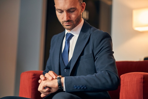 Knappe jongeman in elegant pak zittend in de fauteuil en zijn pols opstekend terwijl hij de tijd controleert