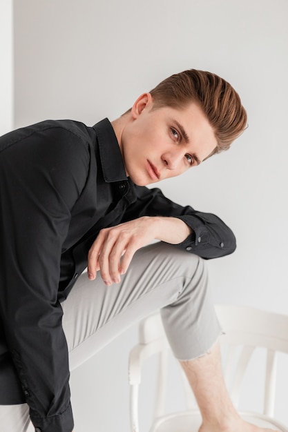 Knappe jongeman in elegant zwart shirt in vintage modieuze broek poseren in de buurt van houten stoel in studio. Aantrekkelijke man mannequin in klassieke stijlvolle kleding in de buurt van witte muur in de kamer.