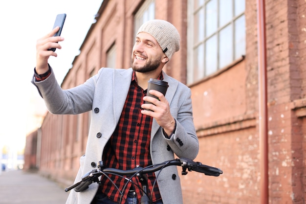 Knappe jongeman in grijze jas en hoed die op straat loopt en koffie drinkt en selfie neemt.