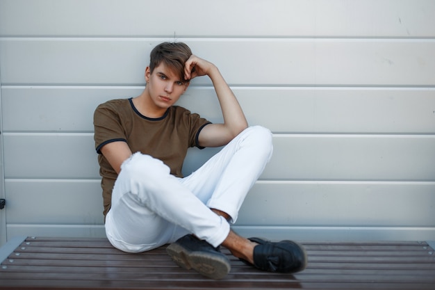 Knappe jongeman in modieuze kleding rust op een bankje in de buurt van een moderne muur