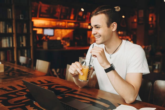 Knappe jongeman met creatieve kapsel in lichte casual shirt aan tafel zitten met laptop en glas met sinaasappelsap te houden