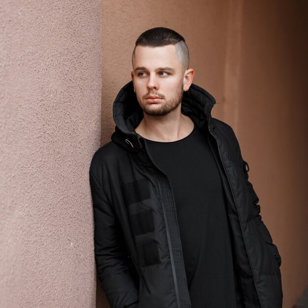 Knappe jongeman met een kapsel in een stijlvolle zwarte warme jas poseren buiten in de buurt van de muur