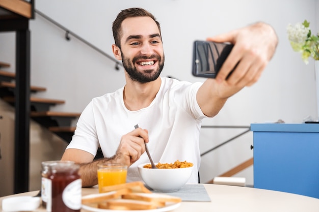 Knappe jongeman ontbijten zittend in de keuken, een selfie nemen