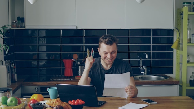 Knappe jongeman ontvangt goed nieuws tijdens het lezen van een papieren brief in de keuken terwijl hij thuis ontbijt