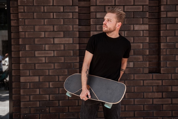 Knappe jongeman skateboarder die tegen bakstenen stadsmuur loopt met een houten longboard en nadenkend naar de grond kijkt, draagt een zwarte outfit en witte sneakers. Denkt na over nieuwe extreme truc