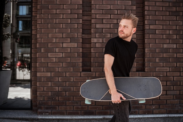 Knappe jongeman skateboarder die tegen bakstenen stadsmuur loopt met een houten longboard en nadenkend naar de grond kijkt, draagt een zwarte outfit en witte sneakers. Denkt na over nieuwe extreme truc