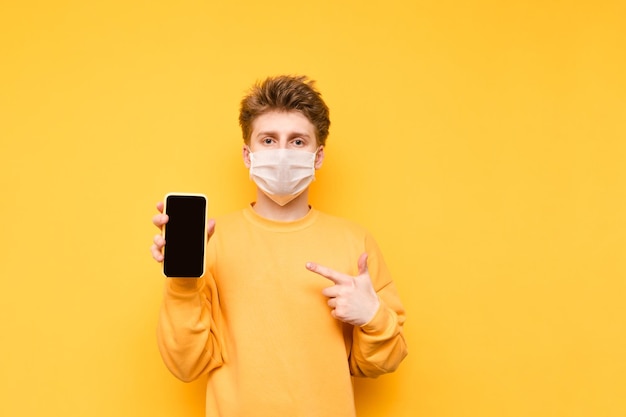 Knappe jongen met een gaasmasker staat met een smartphone in zijn hand op een gele achtergrond