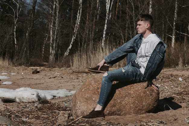 Knappe jongen zittend op de steen in de buurt van de bosweg in het voorjaar en kijken naar de horizon.