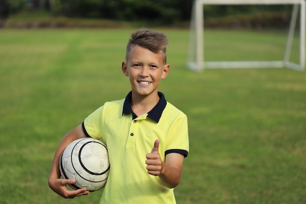 Knappe jongensvoetballer in een geel t-shirt houdt de bal in zijn hand. tweede hand verschijnt duimen