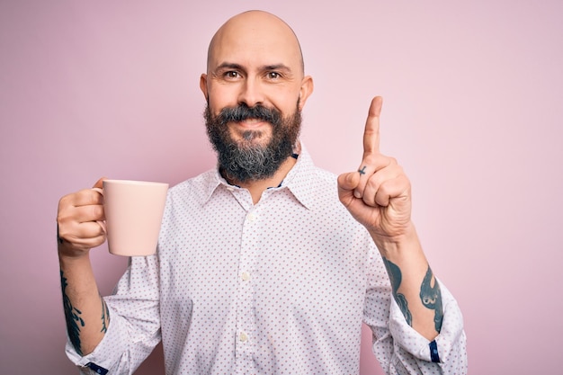 Knappe kale man met baard en tatoeage kopje koffie drinken over roze achtergrond verrast met een idee of vraag wijzende vinger met blij gezicht nummer één