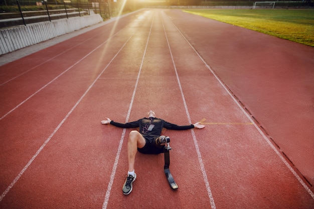 Knappe kaukasische gehandicapte sportieve jongeman gekleed in sportkleding en met kunstbeen liggend op het circuit en muziek luisteren via slimme telefoon.