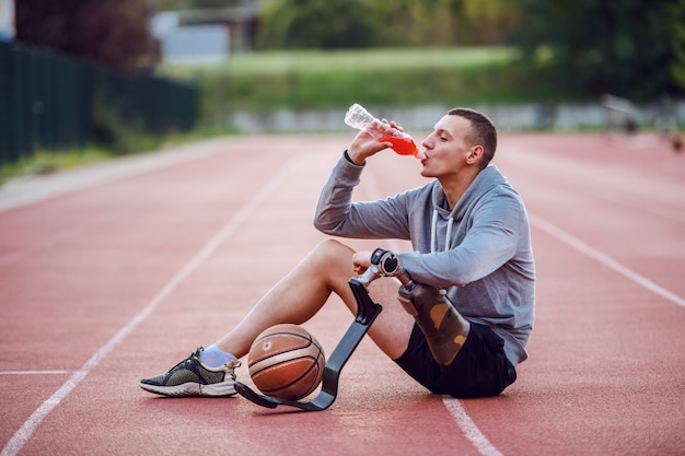 Knappe kaukasische sportieve gehandicapte man in sportkleding zittend op het circuit en verfrissing drinken. tussen de benen zit een basketbalbal.