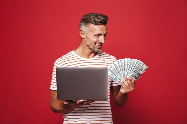 Knappe kerel in gestreept t-shirt glimlachend terwijl hij een fan van geld contant geld en een laptop vasthoudt die op rood wordt geïsoleerd?