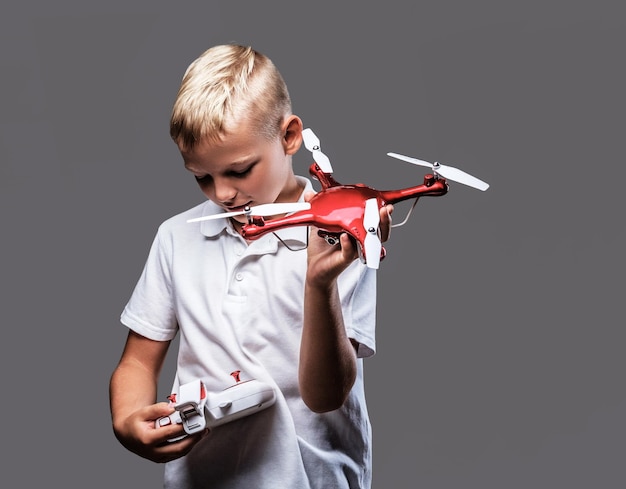 Knappe kleine jongensbokser met blond haar gekleed in een wit t-shirt heeft een quadcopter en afstandsbediening. Geïsoleerd op een grijze achtergrond.