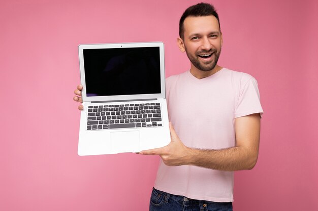 Knappe lachende brunet man met laptopcomputer kijken camera in tshirt op geïsoleerde roze