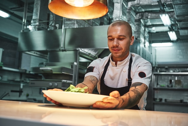 knappe lachende chef-kok met tatoeages in zwarte schort met klaar gerecht in moderne restaurantkeuken