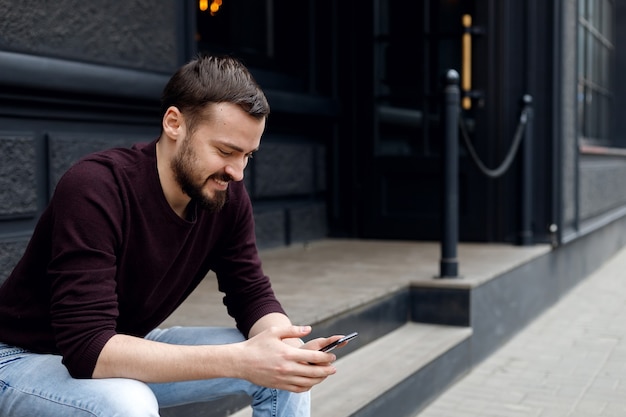 Knappe lachende jongeman die mobiele telefoon gebruikt terwijl hij buiten op trappen zit