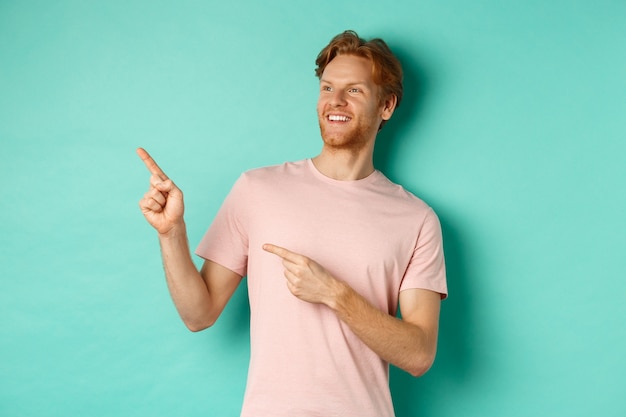 Knappe lachende man met rood haar en baard die opgetogen kijkt, wijzend naar de banner in de linkerbovenhoek, staande in t-shirt over mint achtergrond.