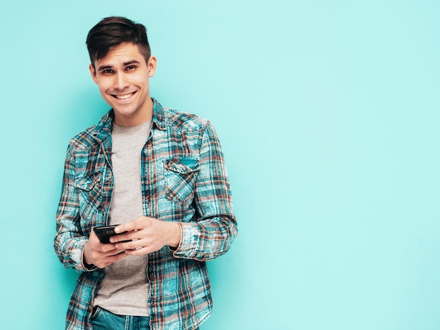 Knappe lachende model Sexy stijlvolle man gekleed in geruit hemd en spijkerbroek Mode hipster man poseren in de buurt van blauw in studio geïsoleerd houden smartphone kijken naar mobiele telefoon scherm met behulp van apps