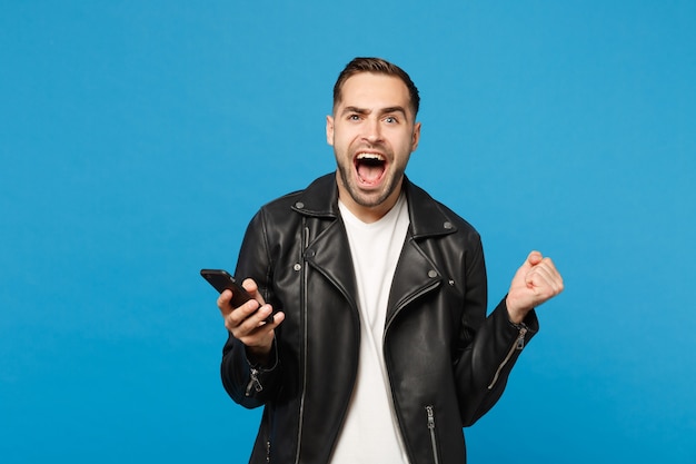 Knappe leuke dolblij opgewonden jonge ongeschoren man in zwart lederen jas wit t-shirt met behulp van mobiele telefoon geïsoleerd op blauwe muur achtergrond studio portret. Mensen levensstijl concept Mock up kopie ruimte