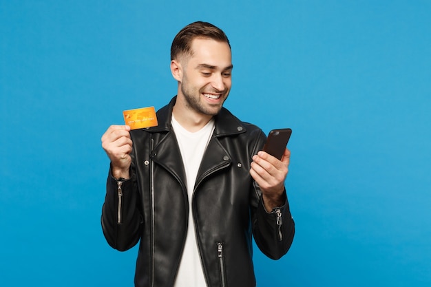 Knappe leuke stijlvolle jonge ongeschoren man in zwarte jas wit t-shirt in de hand houden mobiele telefoon, creditcard geïsoleerd op blauwe muur achtergrond studio portret. Mensen levensstijl concept. Bespotten kopie ruimte