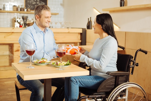 Knappe liefdevolle blonde man die lacht en een cadeau geeft aan zijn aantrekkelijke glimlachende gehandicapte vrouw tijdens een romantisch diner