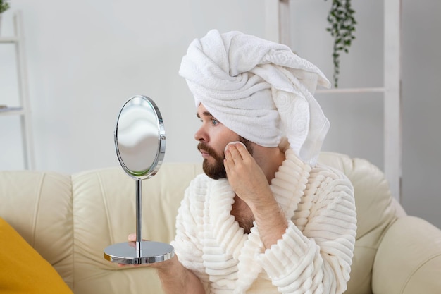 Knappe man die de gezichtshuid schoonmaakt met wattenschijfjes en naar de spiegel kijkt. Spa-, lichaams- en huidverzorging voor mannelijk concept.