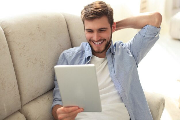 Knappe man gebruikt een digitale tablet en glimlacht terwijl hij thuis op de bank rust.