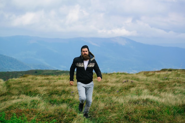 Knappe man hipster met baard en snor in warme jas en hoed draait op de bergtop op zonnige zomerdag op bewolkte hemel