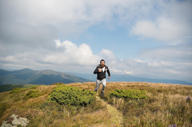 Knappe man hipster of kerel loper met baard en snor in hoed zonnige buiten lopen op bergtop met groen gras tegen bewolkte hemel op natuurlijke achtergrond