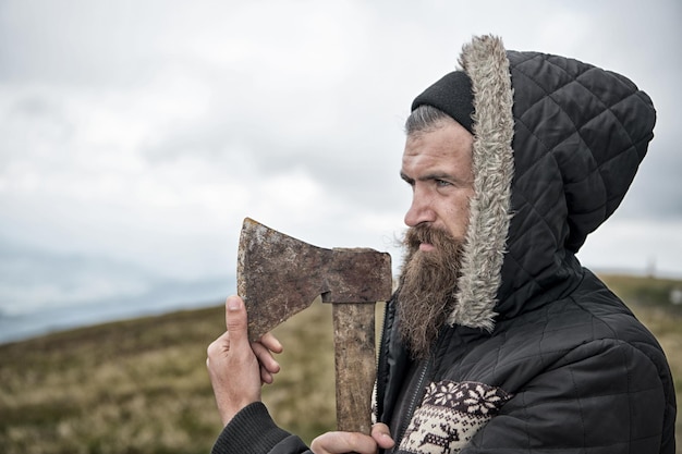 Knappe man hipster of man met baard en snor op ernstig gezicht in hoed en jas houdt roestige bijl met houten gevest buiten op bergtop tegen bewolkte hemel op natuurlijke achtergrond, kopieer ruimte