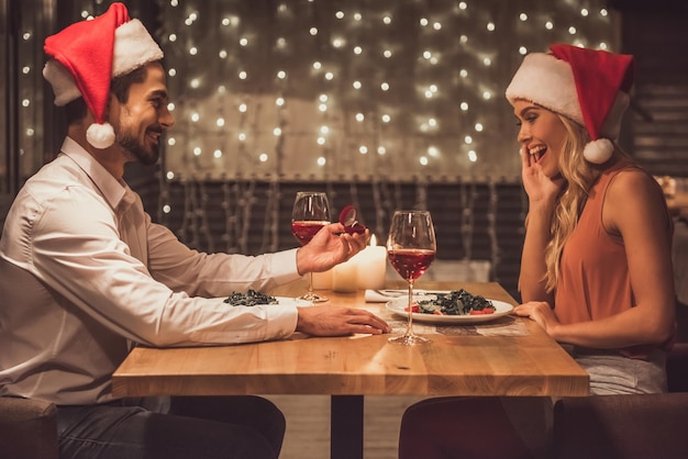 Knappe man houdt een verlovingsring vast en stelt zijn vriendin voor terwijl ze gekleed zijn in kerstmutsen die Nieuwjaar vieren in een restaurant