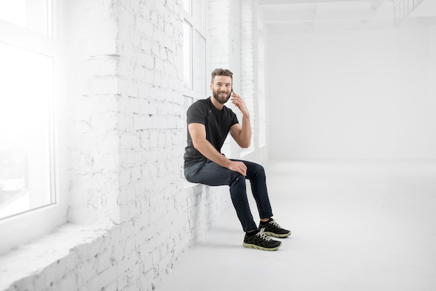 Knappe man in de zwarte sportkleding praten met telefoon zittend op het raam in het interieur van de witte sportschool