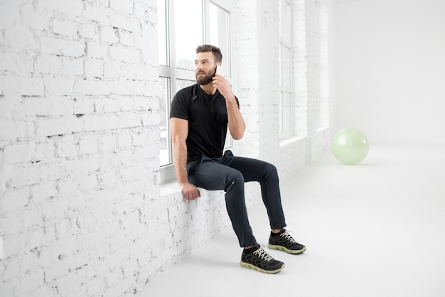 Knappe man in de zwarte sportkleding praten met telefoon zittend op het raam in het interieur van de witte sportschool