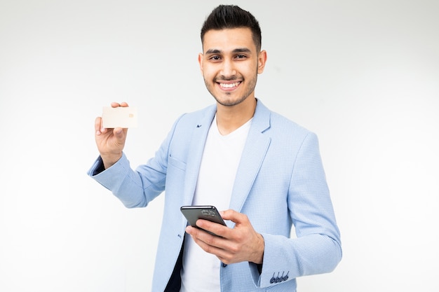 Knappe man in een blauwe jas met een creditcard met een mockup en een telefoon in zijn hand op een witte studio achtergrond