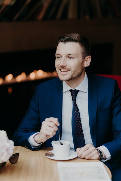 Knappe man in formele kleding, heeft vrolijke uitdrukking, heeft dinerpauze in cafetaria, drinkt aromatische koffie