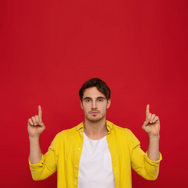 knappe man in gele overhemd wijzend met vingers omhoog geïsoleerd op rode achtergrond