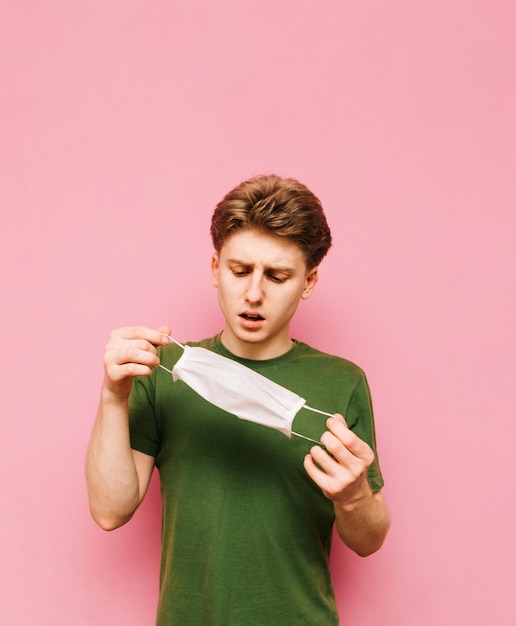 Knappe man in groene tshirt staat op een roze achtergrond met een medisch masker in zijn handen